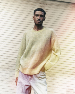 Editorial portrait of model Albhei Gazi wearing a light knit sweater and faded jeans, standing against a cream-colored textured wall. 