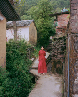 Ngawang Jinpa wearing a red embroidered dress, standing on a narrow village pathway surrounded by stone walls and greenery, for the Eka campaign shot by photographer Tenzin Lhagyal for ALF. 