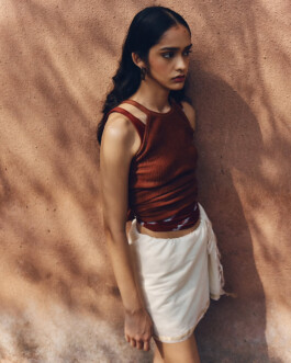 Editorial portrait of model Theia Maeve wearing a rust-colored sleeveless top and white wrap skirt against a textured wall for the Maatii Studio campaign. 