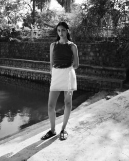 Black-and-white outdoor fashion portrait of model Theia Maeve standing beside water in a dark sleeveless top and white skirt for the Maatii Studio campaign 