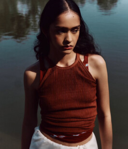 Editorial portrait of model Theia Maeve wearing a rust brown textured top beside water for the Maatii Studio campaign. 