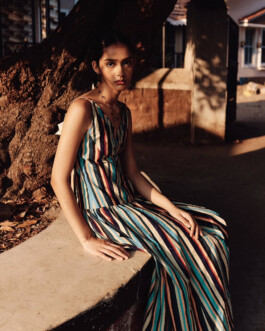 Model Theia Maeve seated outdoors in warm golden light wearing a multicolored striped dress for the Maatii Studio campaign. 