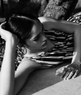 Black and white editorial image of model Theia Maeve reclining outdoors in a striped dress for the Maatii Studio campaign. 